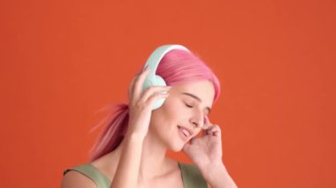 Pembe saçlı memnun bir genç kadın stüdyodaki turuncu duvarın üzerinde duran kulaklıkla müzik dinliyor.