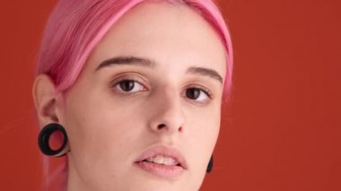 Pembe saçlı hoş bir genç kadının yakın çekim görüntüsü kameraya bakıyor. Stüdyonun turuncu duvarının üzerinde duran aynaya bakıyor.