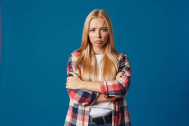 Sarışın, hoşnutsuz, kolları çapraz pozlar veren genç bir kadın mavi arka planda izole edilmiş.