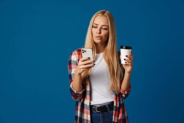 Kafası karışmış güzel sarışın kız mavi arka planda izole kahve içerken akıllı telefon kullanıyor.