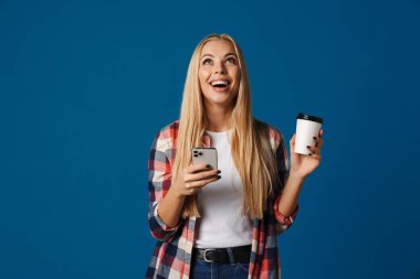 Neşeli güzel sarışın kız mavi arka planda izole edilmiş kahve içerken akıllı telefon kullanıyor.