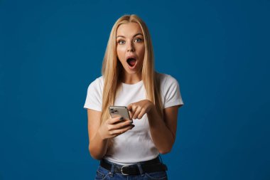 Sarışın beyaz kadın mavi arka planda izole edilmiş cep telefonu kullanırken haykırıyor.