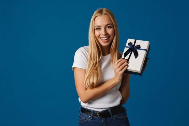 Mavi arka planda saklanmış hediye kutusuyla poz verirken gülümseyen mutlu güzel kız.