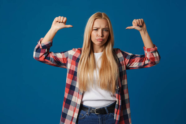 Blonde young displeased woman pointing fingers at herself isolated over blue background