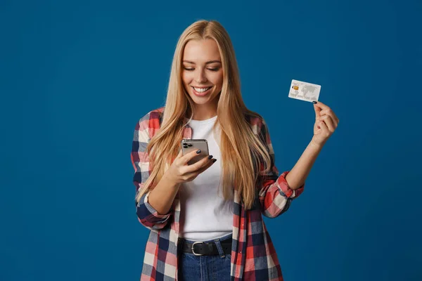 ブロンド幸せな女性を使用して携帯電話と示すクレジットカード隔離された青の背景 — ストック写真