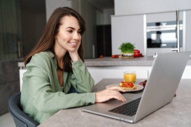 Evde mutfakta otururken dizüstü bilgisayarda çalışan çekici genç bir kadın.