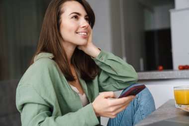 Mutfakta cep telefonu kullanırken gülümseyen neşeli, güzel bir kadın.