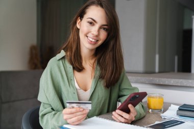 Evraklarla uğraşırken cep telefonu ve kredi kartı kullanan mutlu bir kadın.