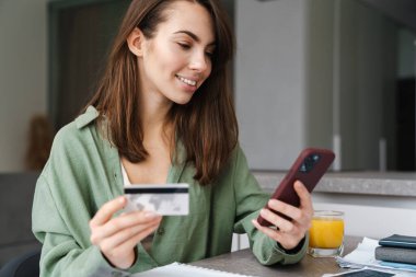 Evde masada otururken cep telefonu ve kredi kartı kullanan gülümseyen bir kadın.