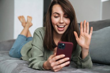 Evdeki kanepede uzanırken el kol hareketi yapan ve cep telefonu kullanan genç bir kadın.