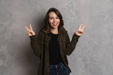 Mutlu gülümseyen kadın portresi gri arka planda tek başına duruyor, barış jesti gösteriyor.