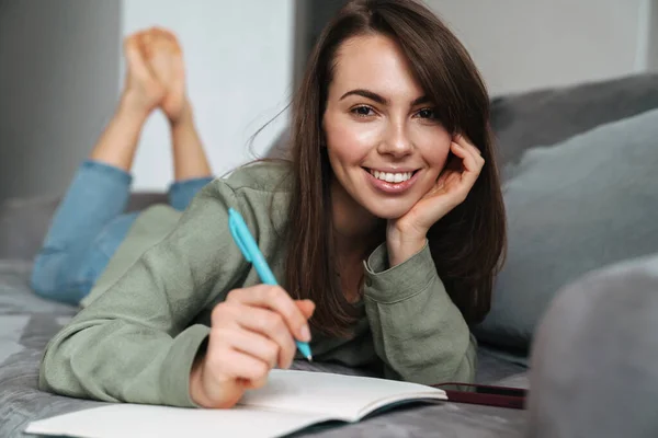 Młoda Uśmiechnięta Kobieta Zapisuje Notatki Leżąc Kanapie Domu — Zdjęcie stockowe