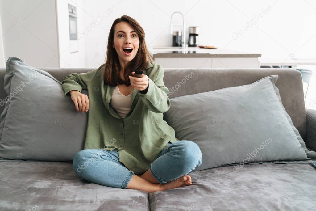 Happy young woman watching tv on sofa, holding remote control