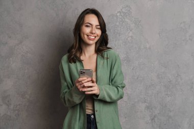 Gri duvarda izole edilmiş cep telefonu kullanırken göz kırpan mutlu esmer kız.