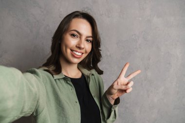 Gri duvardan izole edilmiş selfie çekerken huzur işareti gösteren mutlu güzel kız.
