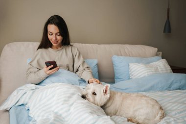 Mutlu beyaz kadın evde yatakta otururken köpeğini okşuyor ve cep telefonu kullanıyor.
