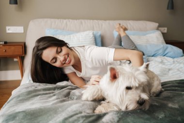 Mutlu beyaz kadın evde yatarken köpeğini okşuyor.