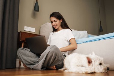 Mutlu beyaz kadın evinde köpeğiyle otururken dizüstü bilgisayar kullanıyor.