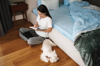 Mutlu genç esmer kadın, yatak odasında modellik yapıyor, yerde köpeğiyle ve dizüstü bilgisayarıyla oturuyor.