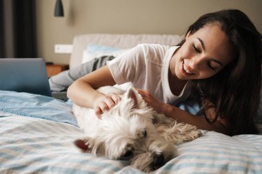 Mutlu beyaz kadın evde yatarken köpeğini okşuyor.