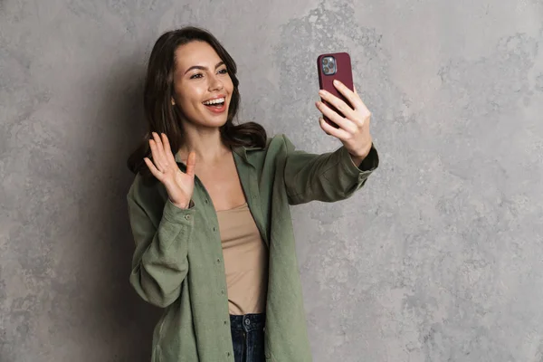 快乐可爱的女孩一边挥手一边用灰色墙壁上的手机自拍 — 图库照片