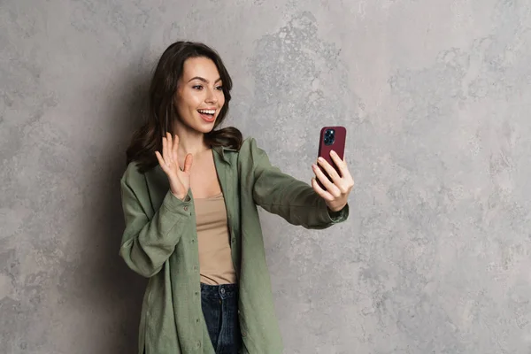 快乐可爱的女孩一边挥手一边用灰色墙壁上的手机自拍 — 图库照片