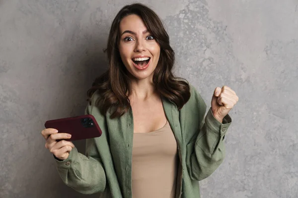 快乐的女孩一边在灰色墙壁外的手机上玩网络游戏 一边做出优胜的姿态 — 图库照片