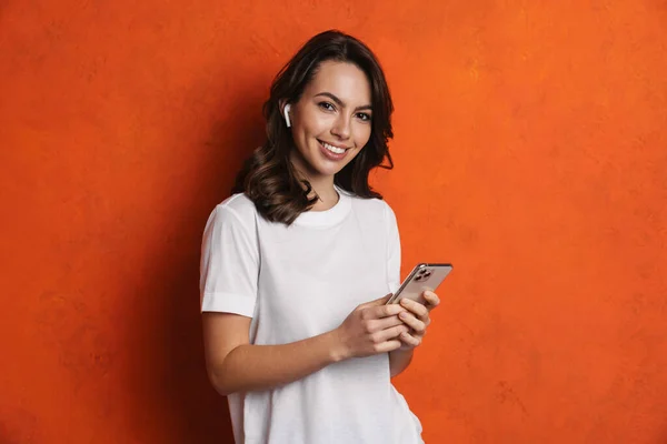 戴耳机的快乐女孩笑着用隔离在橙色墙壁上的手机 — 图库照片