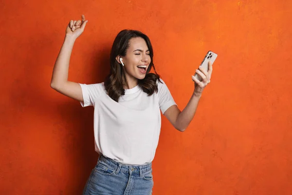 戴耳机的快乐女孩一边唱歌一边跳舞一边用隔离在橙色墙上的手机 — 图库照片