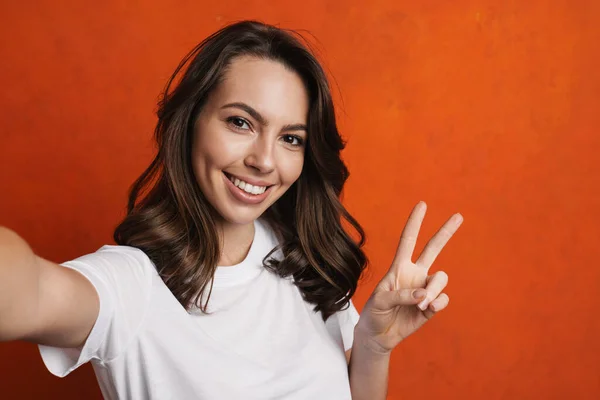 快乐美丽的女孩一边举手投足和平标志一边在橙色墙上拍自拍照 — 图库照片