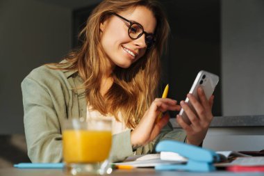 Evde ödev yaparken cep telefonu kullanan mutlu, hoş bir bayan öğrenci.