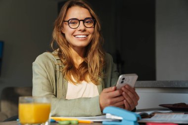 Evde ödev yaparken cep telefonu kullanan mutlu, hoş bir bayan öğrenci.