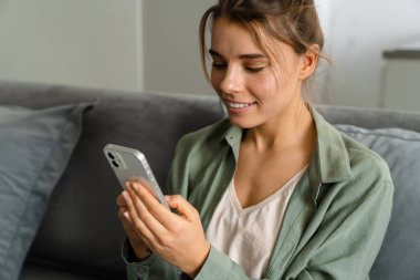 Mutlu güzel bir kadın evdeki koltukta otururken cep telefonu kullanıyor.