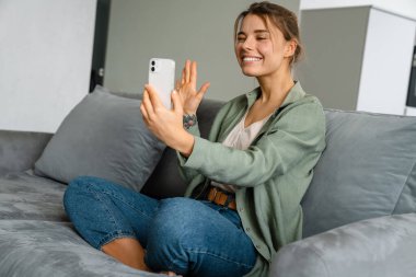 Heyecanlı güzel bir kadın evde cep telefonuyla selfie çekerken el sallıyor.