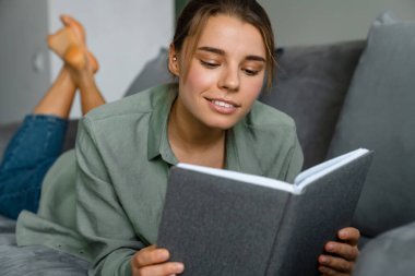 Evdeki kanepede uzanmış kitap okuyan güzel bir kadın.