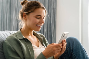 Mutlu güzel bir kadın evdeki koltukta otururken cep telefonu kullanıyor.