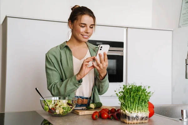 Feliz Mulher Agradável Usando Telefone Celular Enquanto Prepara Salada Casa — Fotografia de Stock