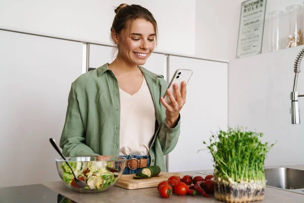Feliz Mujer Agradable Usando Teléfono Móvil Mientras Prepara Ensalada Cocina —  Fotos de Stock