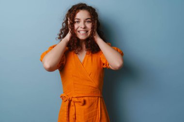 Mutlu kızıl saçlı kıvırcık kız gülümsüyor ve kulaklarını kapatıyor mavi arka planda.