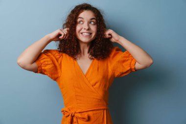 Gülümseyen ve kulaklarını tıkayan mutlu kızıl saçlı kız mavi arka planda izole edilmiş.