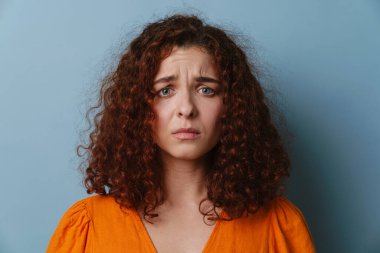 Mutsuz kızıl kadın yüzünü buruşturuyor ve mavi arka planda izole edilmiş kameraya bakıyor.