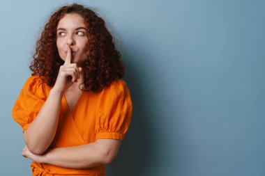 Mutlu kızıl saçlı kıvırcık kız mavi arka planda izole edilmiş sessizlik jesti yapıyor.