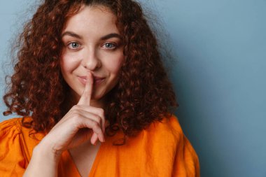 Mutlu kızıl kadın gülümserken mavi arka planda izole edilmiş sessizlik hareketi gösteriyor.