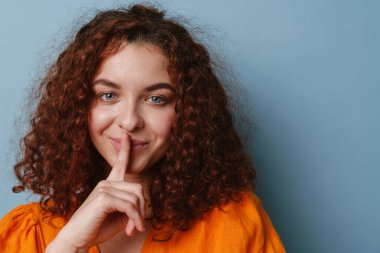 Mutlu kızıl kadın gülümserken mavi arka planda izole edilmiş sessizlik hareketi gösteriyor.