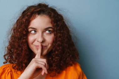 Mutlu kızıl kadın gülümserken mavi arka planda izole edilmiş sessizlik hareketi gösteriyor.