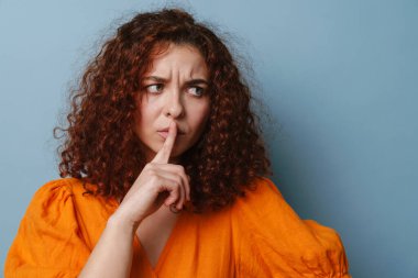 Hoşnutsuz genç kadın, mavi arka planda izole edilmiş sessizlik jesti gösteriyor.