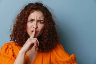 Hoşnutsuz genç kadın, mavi arka planda izole edilmiş sessizlik jesti gösteriyor.