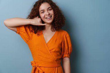 Mutlu kızıl saçlı kıvırcık kız mavi arka planda izole bir şekilde el hareketi ve gülümseme gösteriyor.