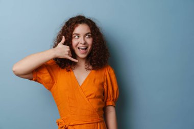 Mutlu kızıl saçlı kıvırcık kız mavi arka planda izole bir şekilde el hareketi ve gülümseme gösteriyor.