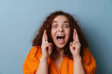 Heyecanlı, kıvırcık saçlı, şans dileyen kız mavi arka planda izole edilmiş.